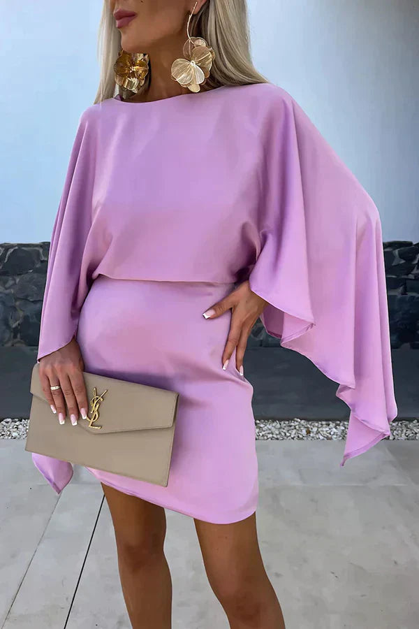 Woman standing in a beige mini dress with cape-inspired sleeves against a plain background.
