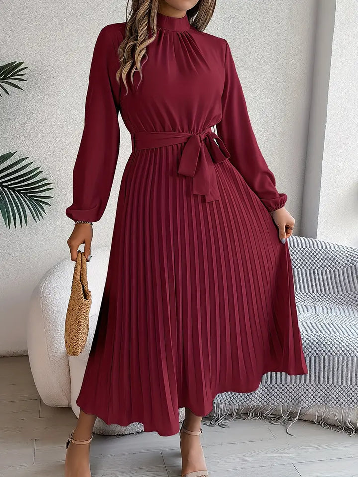 Woman posing in a belted long dress with full sleeves on a plain background.
