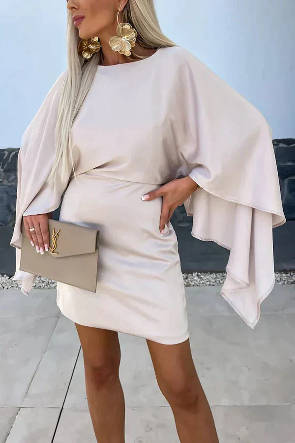Woman posing in a fitted mini dress with flowing batwing cape sleeves on a plain backdrop.