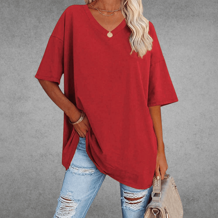 Woman in casual oversized top, holding bag, gray backdrop.