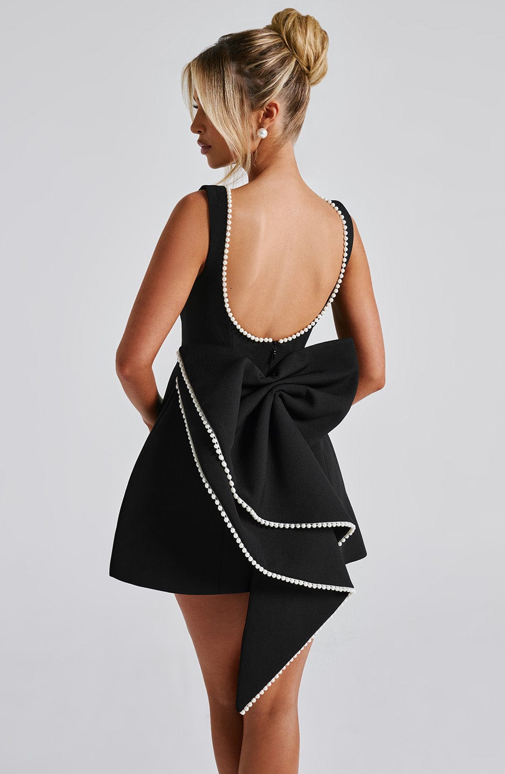 Female model wearing polished mini dress, back pose, gray backdrop.