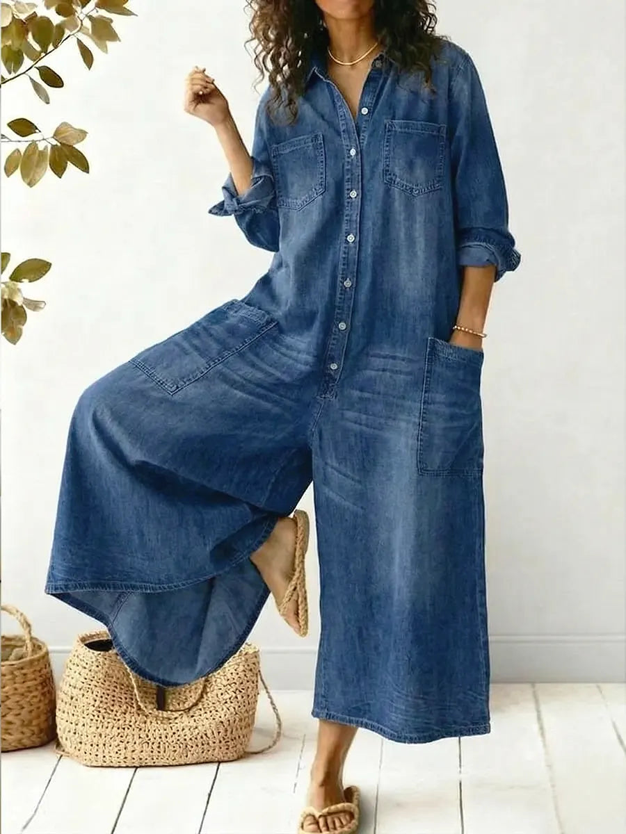 Female model wearing a relaxed blue denim wide-leg jumpsuit on a plain background.