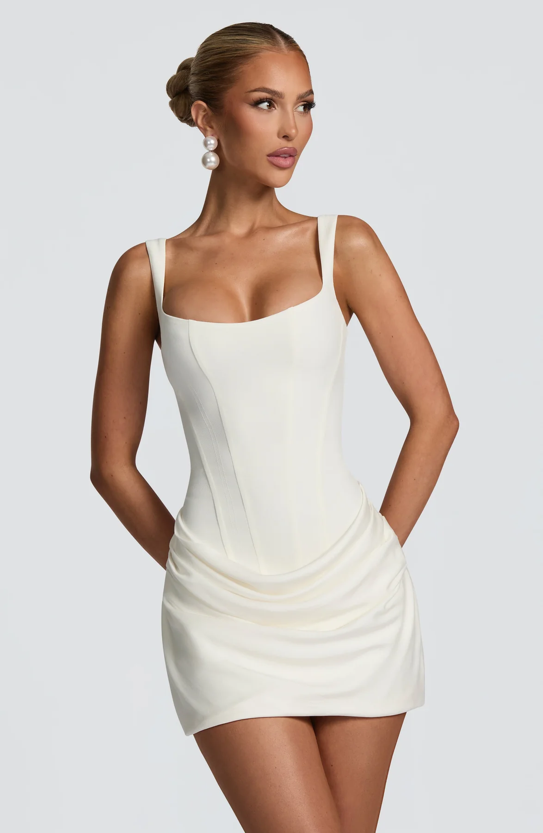 Female model wearing a square neck mini dress with a fitted bodice and draped skirt on a plain background.