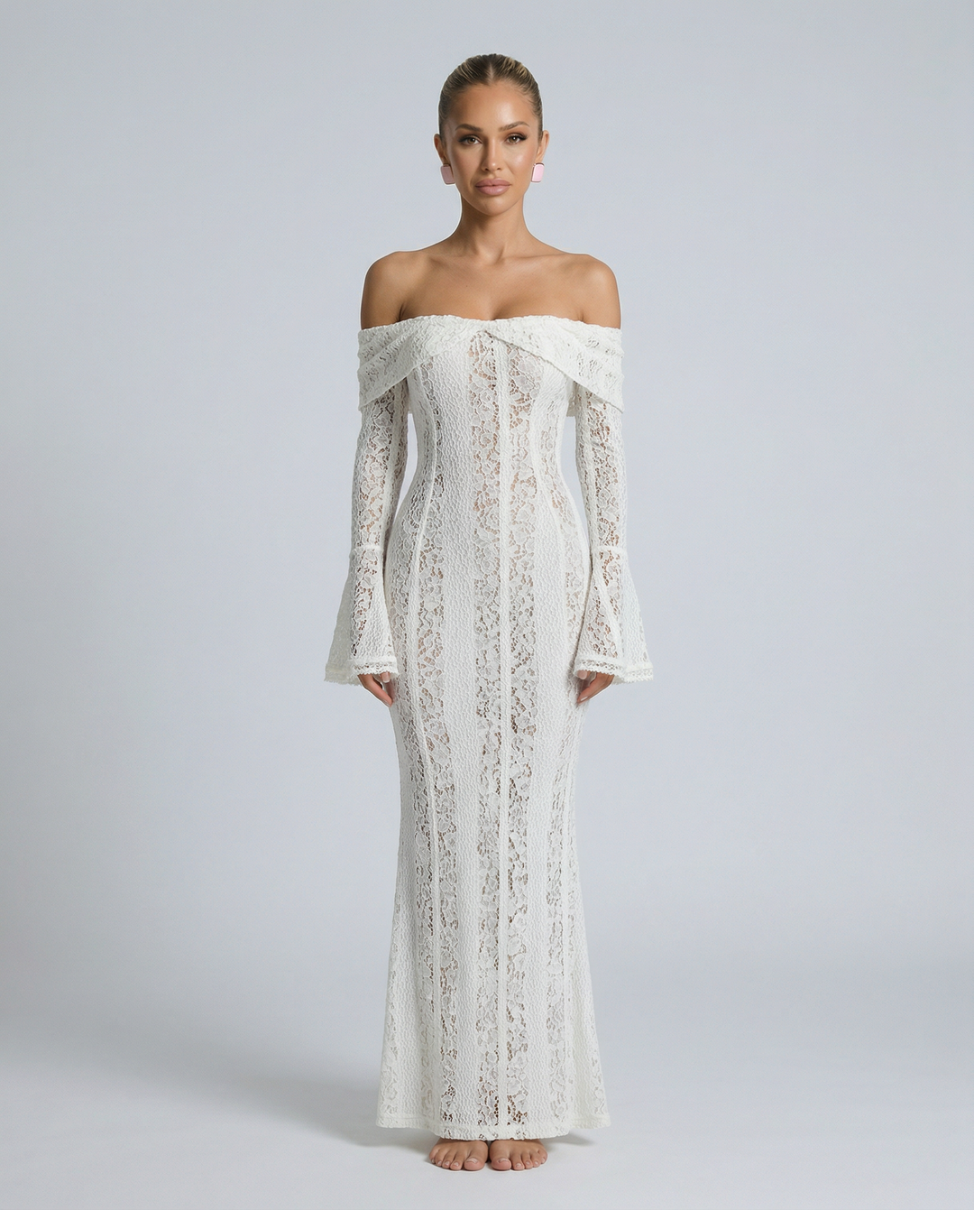 Woman in a white fitted off shoulder maxi dress with long flared sleeves on a plain background.