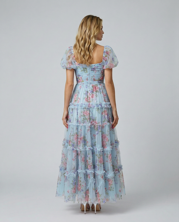 Woman in a blue floral layered maxi dress with square neckline on a plain background.