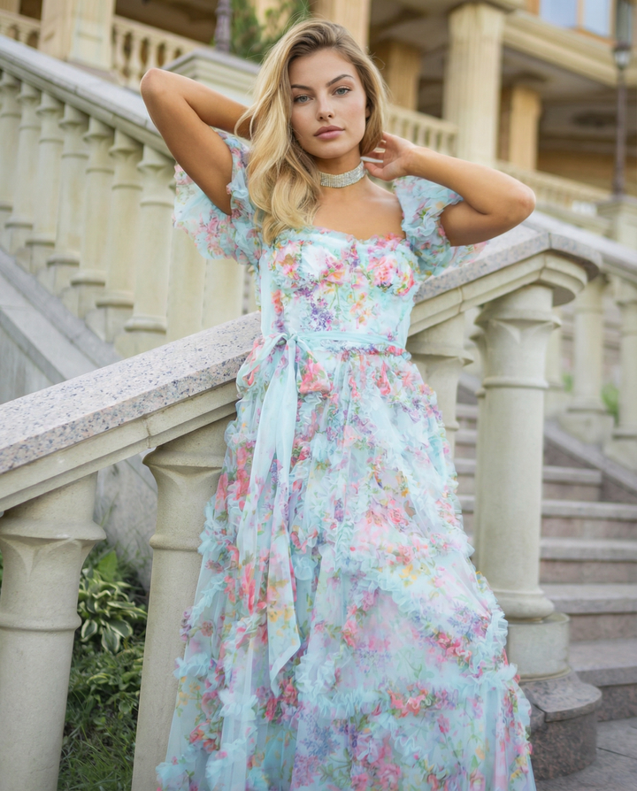 Female model in a flowing floral occasion dress with defined waist on a staircase background.
