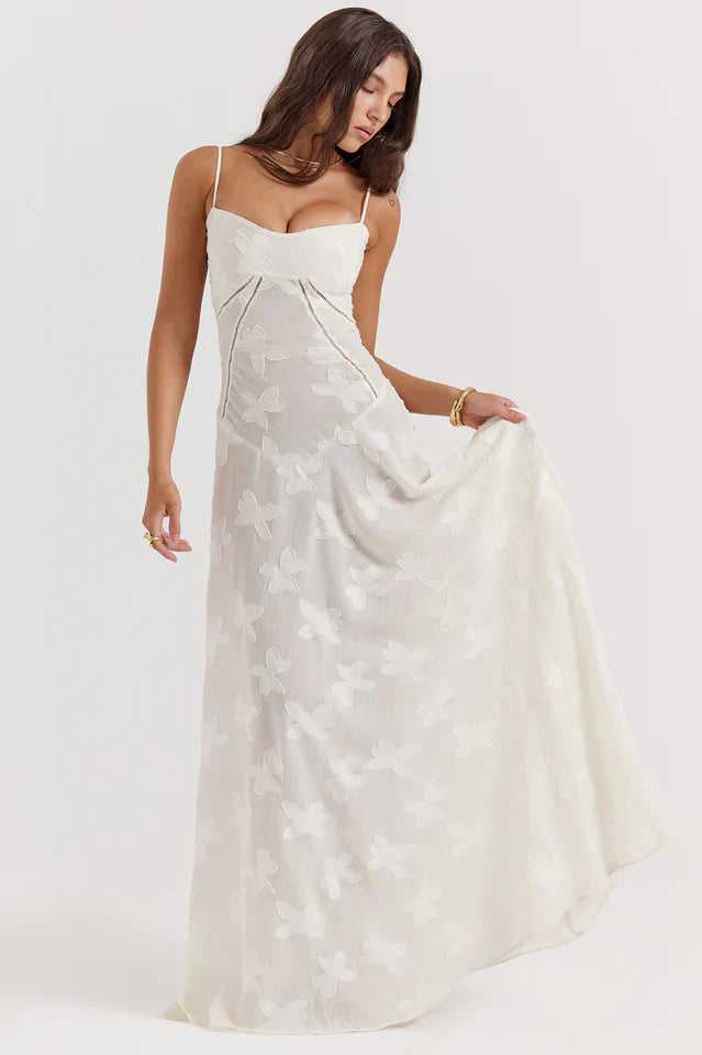 Model in a flowing maxi dress with slim straps and floral print on a plain background.