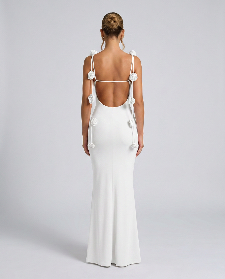 Woman in a white open back maxi dress with floral strap details on a plain background.