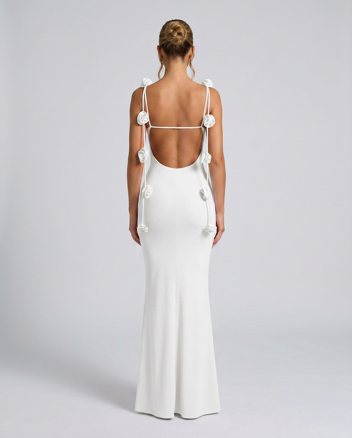Woman in a white open back maxi dress with floral strap details on a plain background.