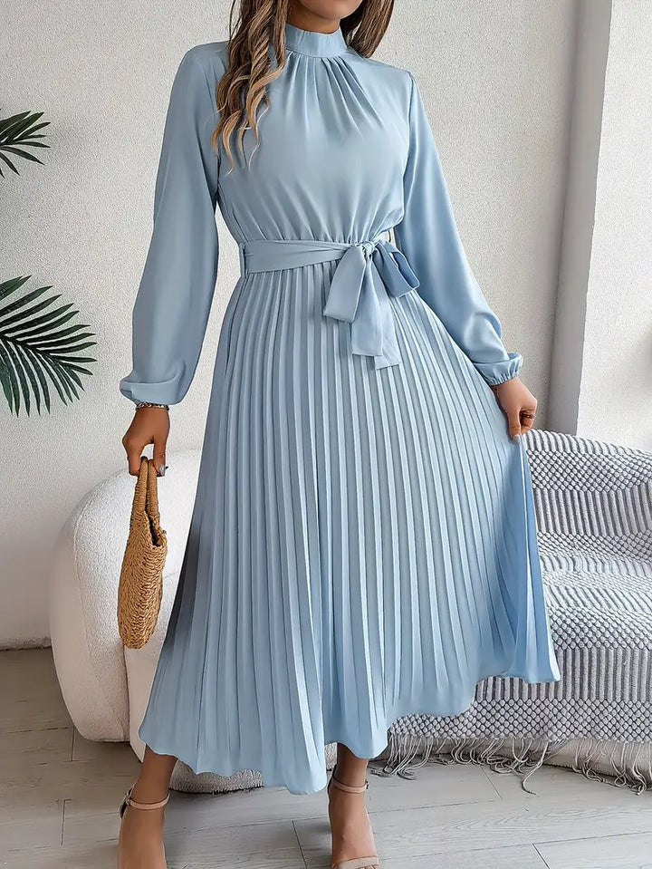 Woman wearing a long sleeve pleated belted dress on a plain background.