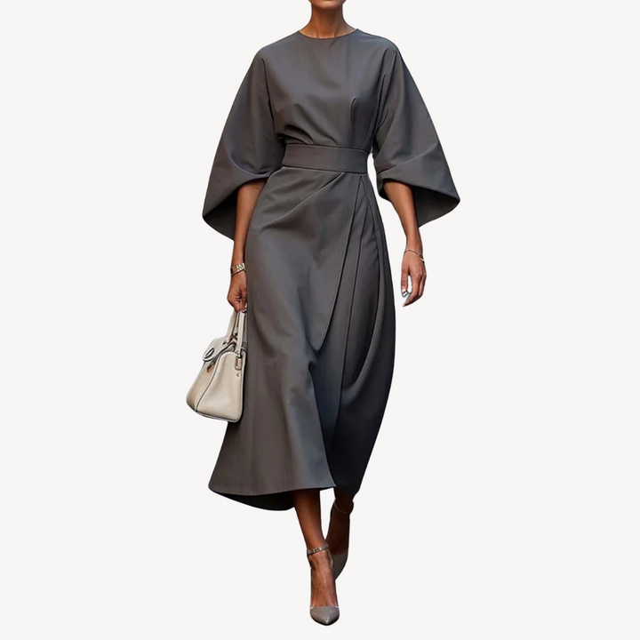 Model in a midi dress with a defined waist and wide sleeves on a plain background.
