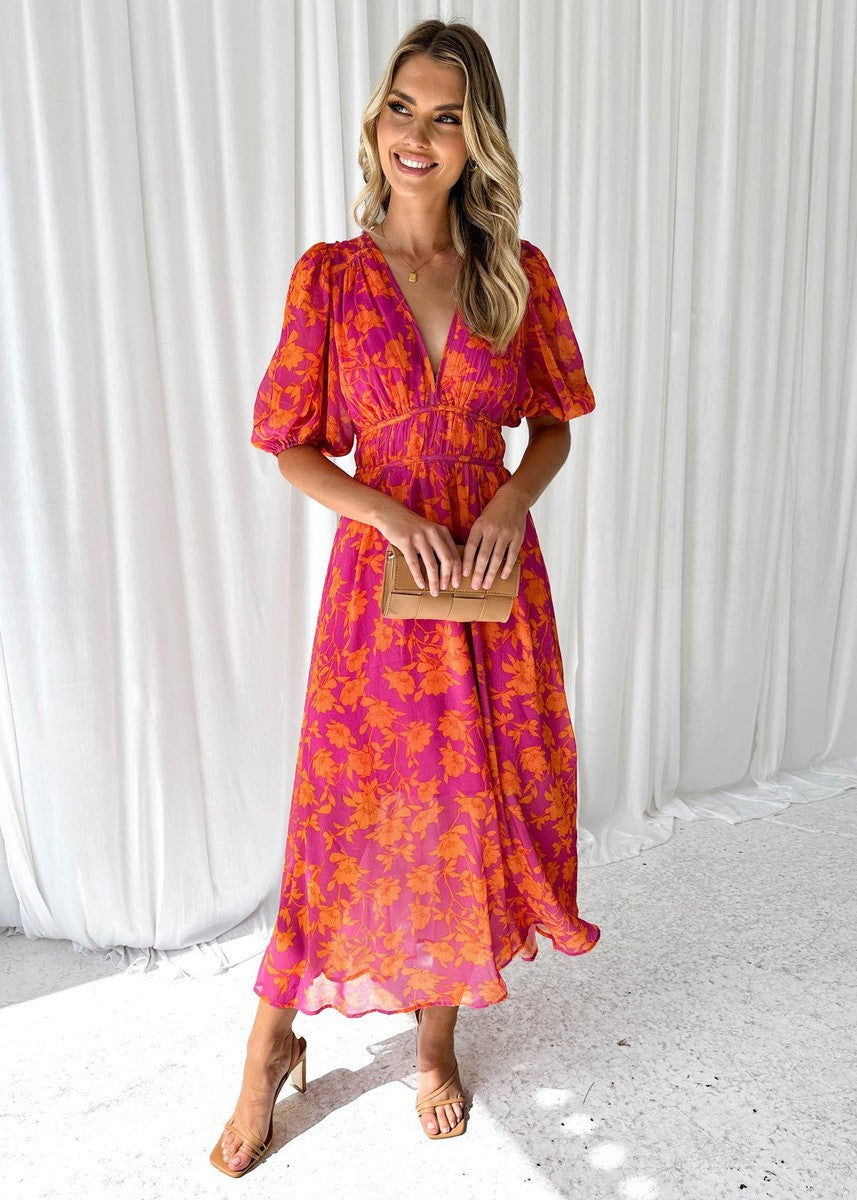Woman showing V-neck floral maxi dress on curtain background, smiling.
