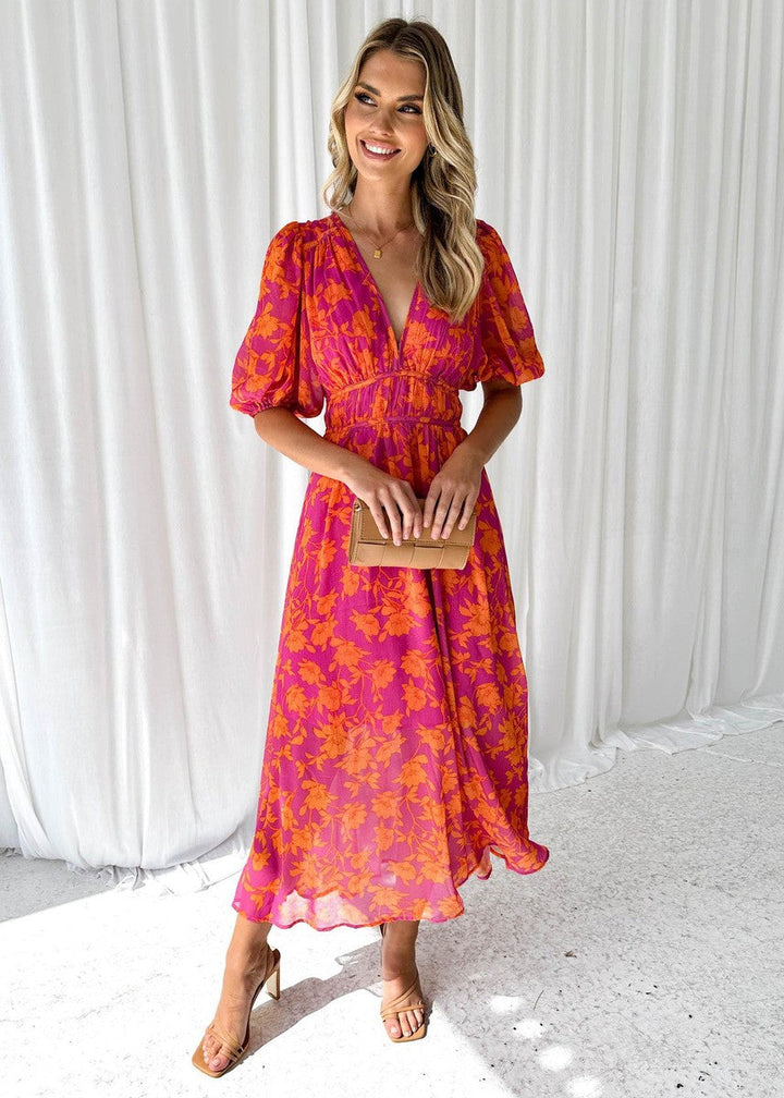 Woman showing V-neck floral maxi dress on curtain background, smiling.
