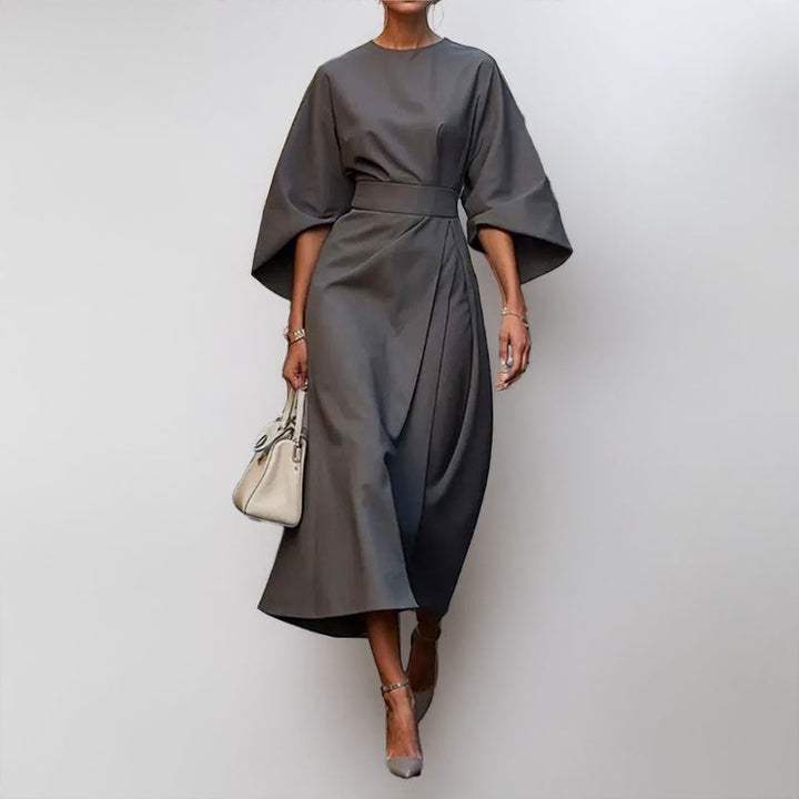 Female wearing an elegant draped maxi dress with satin-like texture on a plain background.